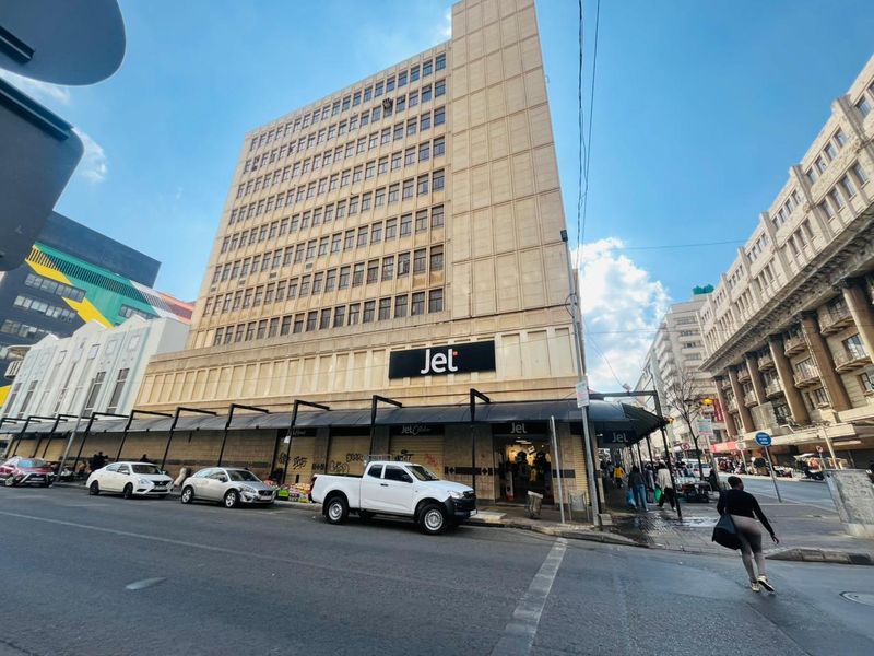 A multi-story commercial building facade featuring a prominent storefront with metal roller shutters and an awning, located in a busy urban street setting.