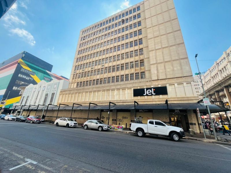 A multi-story commercial building facade featuring ground-level retail space with metal roller shutters and prominent signage in a busy urban street setting.