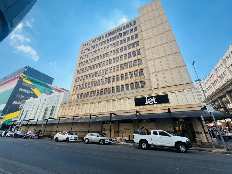 A multi-story commercial building featuring ground-level retail storefronts with metal security shutters and a prominent street-facing facade in an urban setting.