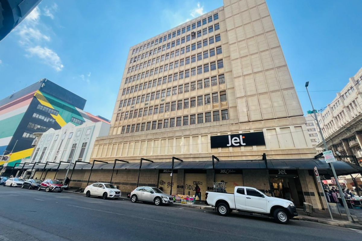 Multi-story commercial building facade featuring ground-level retail storefronts with metal roller shutters and prominent signage in a busy urban street setting.