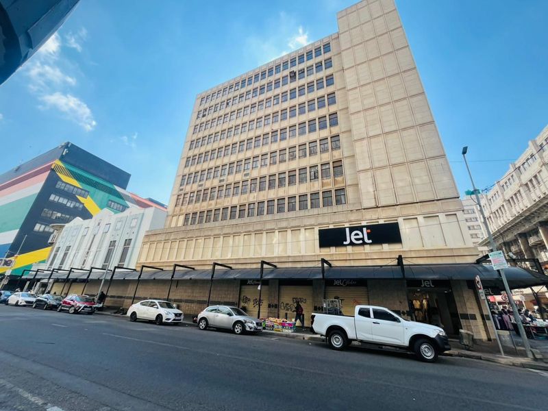 Multi-story commercial building facade featuring ground-level retail storefronts with metal roller shutters and prominent signage in a busy urban street setting.