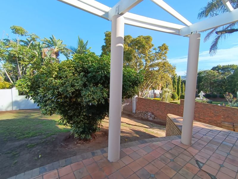 A covered patio area featuring tiled flooring and structural pillars, overlooking a garden with brick boundary walls and established trees.
