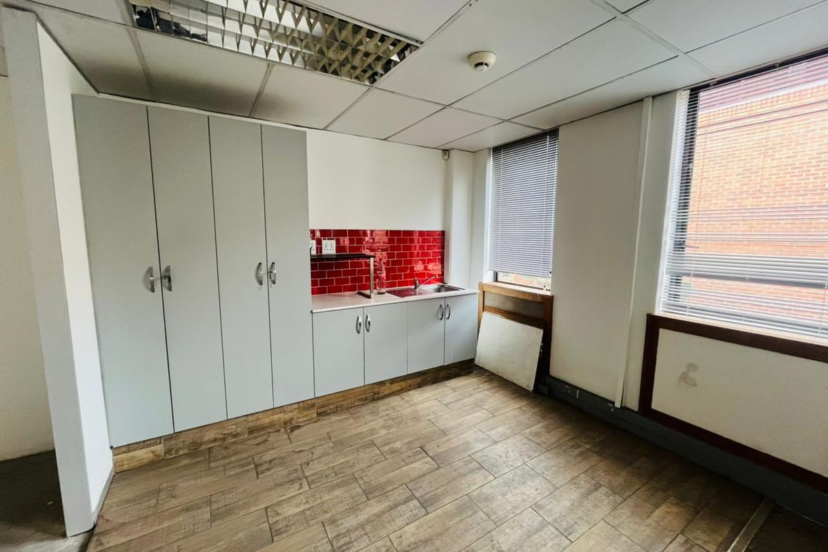 A functional commercial kitchenette featuring built-in cabinetry, a stainless steel sink, red tiled splashback, and wood-look flooring in a professional office setting.