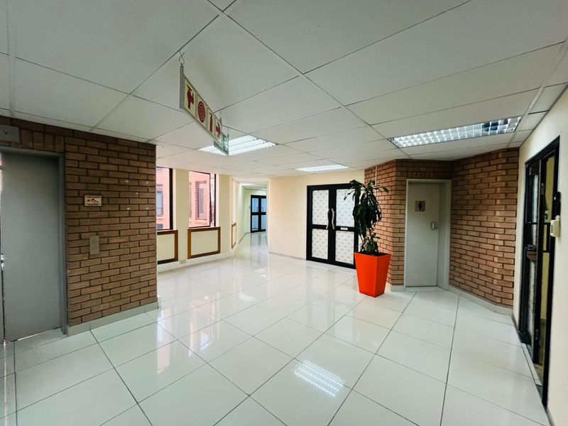 A spacious commercial hallway featuring exposed brick accents, polished tile flooring, and a suspended ceiling with integrated lighting.