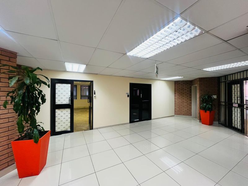 A bright commercial entrance lobby featuring tiled flooring, suspended ceilings with recessed lighting, and exposed brick accents leading to internal office spaces.
