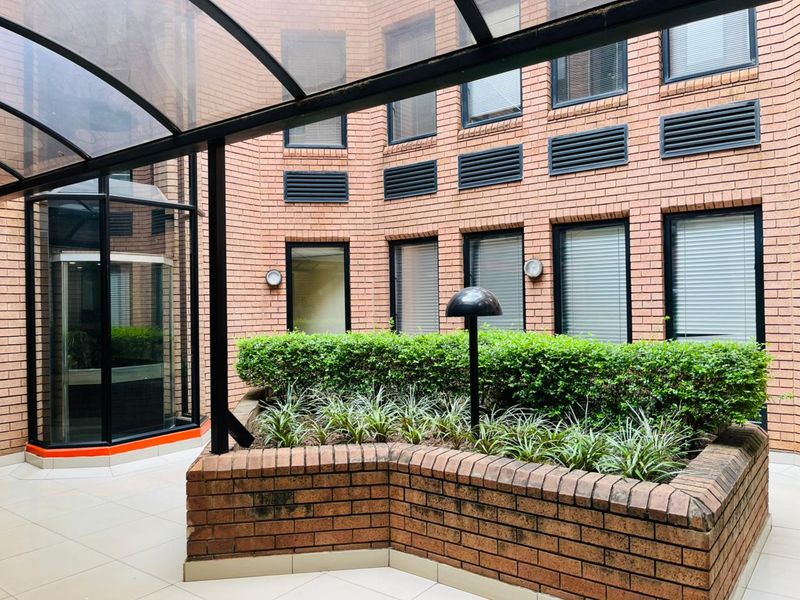 A covered outdoor courtyard area featuring a brick planter box, tiled flooring, and a modern polycarbonate awning, adjacent to a glass elevator shaft.