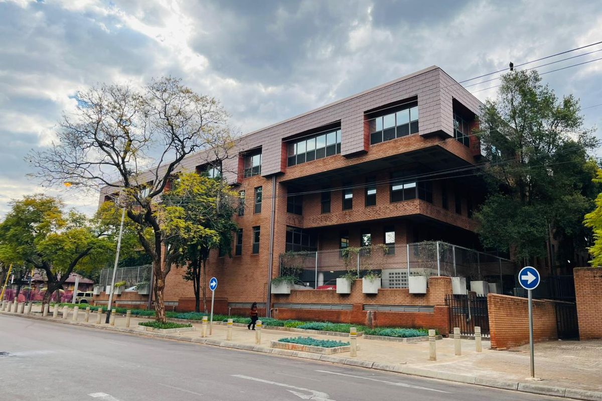 A multi-story commercial building featuring a brick facade, modern tiled roof design, and secure perimeter fencing, situated along a paved street.