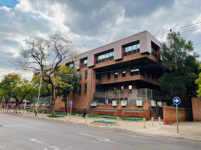 A multi-story commercial building featuring a brick facade, modern tiled roof design, and secure perimeter fencing, situated along a paved street.