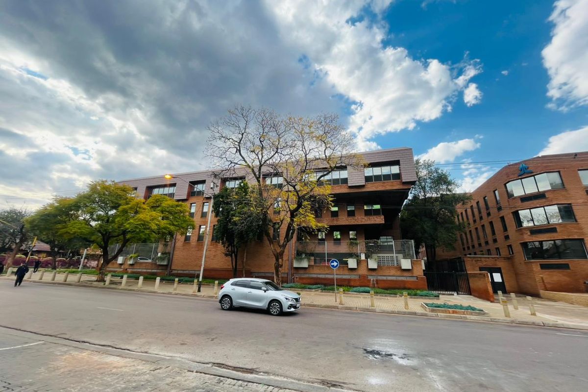 A multi-story commercial brick building featuring large windows and a tiled roof, situated along a paved street with perimeter fencing.