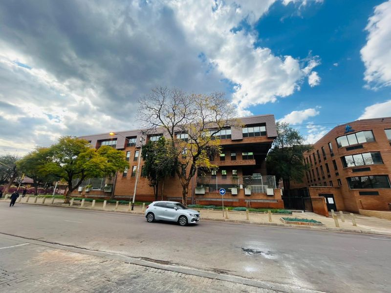A multi-story commercial brick building featuring large windows and a tiled roof, situated along a paved street with perimeter fencing.