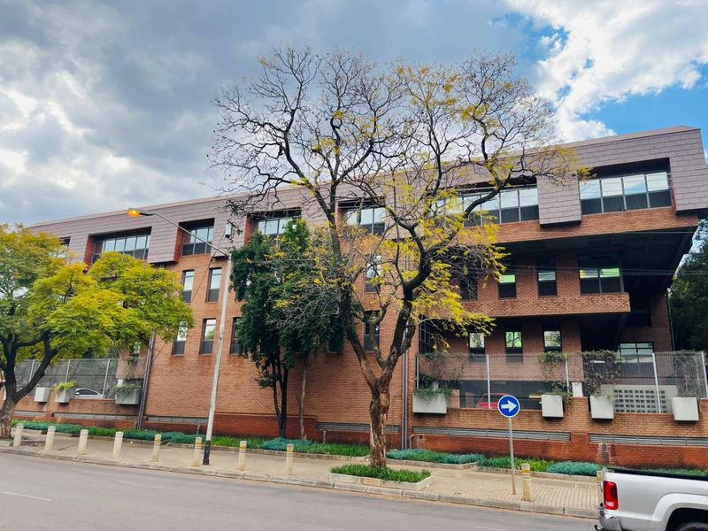 A multi-story commercial building featuring a classic brick facade, large windows, and secure perimeter fencing, situated along a paved street.