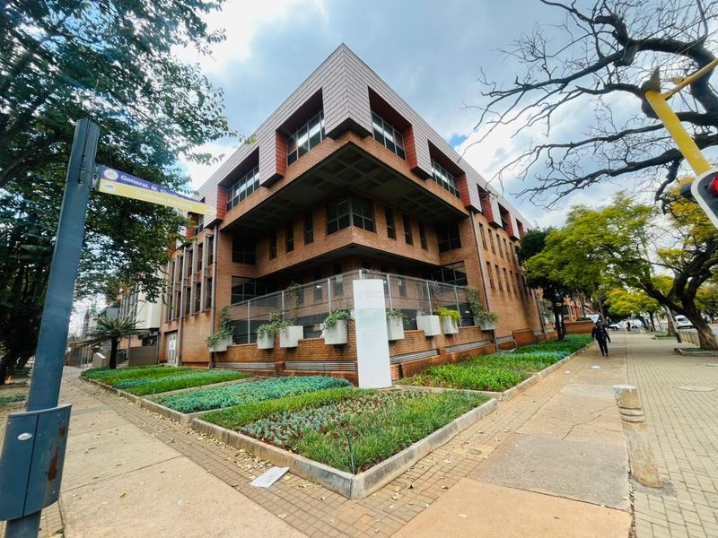 A multi-story commercial office building featuring a brick facade, modern metal window detailing, and landscaped concrete planters along the street-facing perimeter.