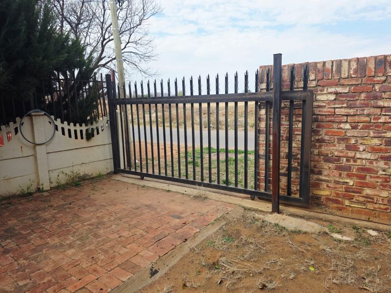 Front entrance area featuring a metal security gate, brick boundary wall, and paved driveway access.