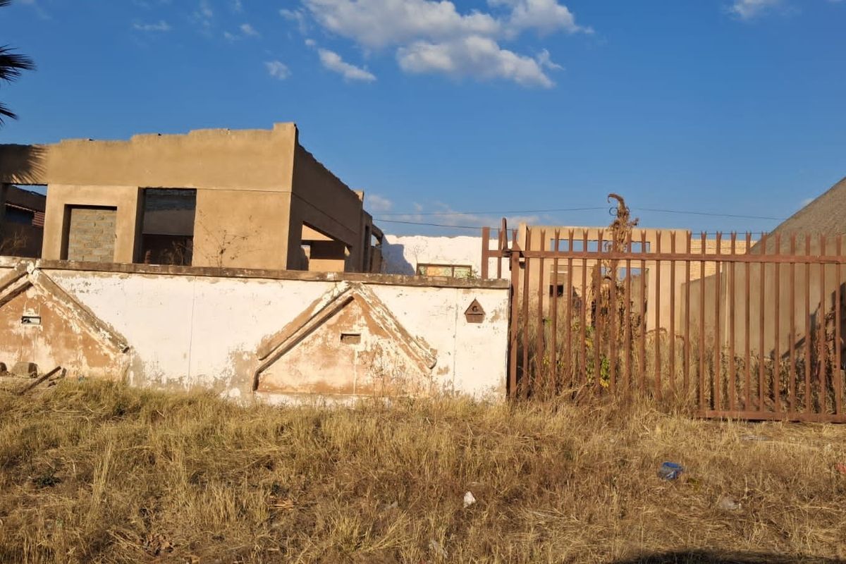 A view of a residential property featuring a dilapidated boundary wall, a rusted metal gate, and an unfinished or severely weathered building structure.