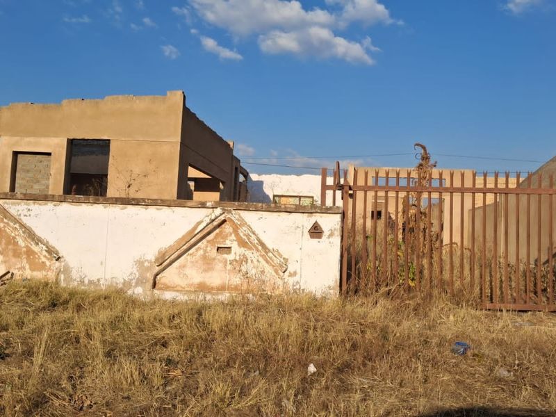 A view of a residential property featuring a dilapidated boundary wall, a rusted metal gate, and an unfinished or severely weathered building structure.
