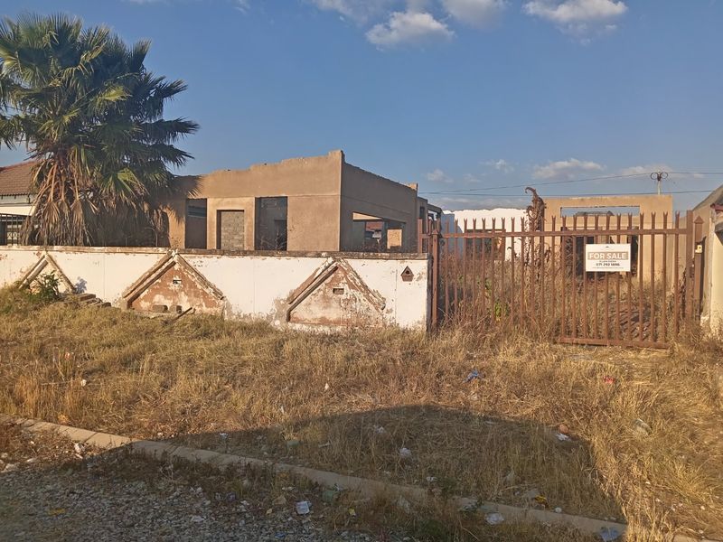 A vacant residential property featuring an unfinished building structure, a perimeter boundary wall, and a metal entrance gate, requiring significant renovation and site clearing.