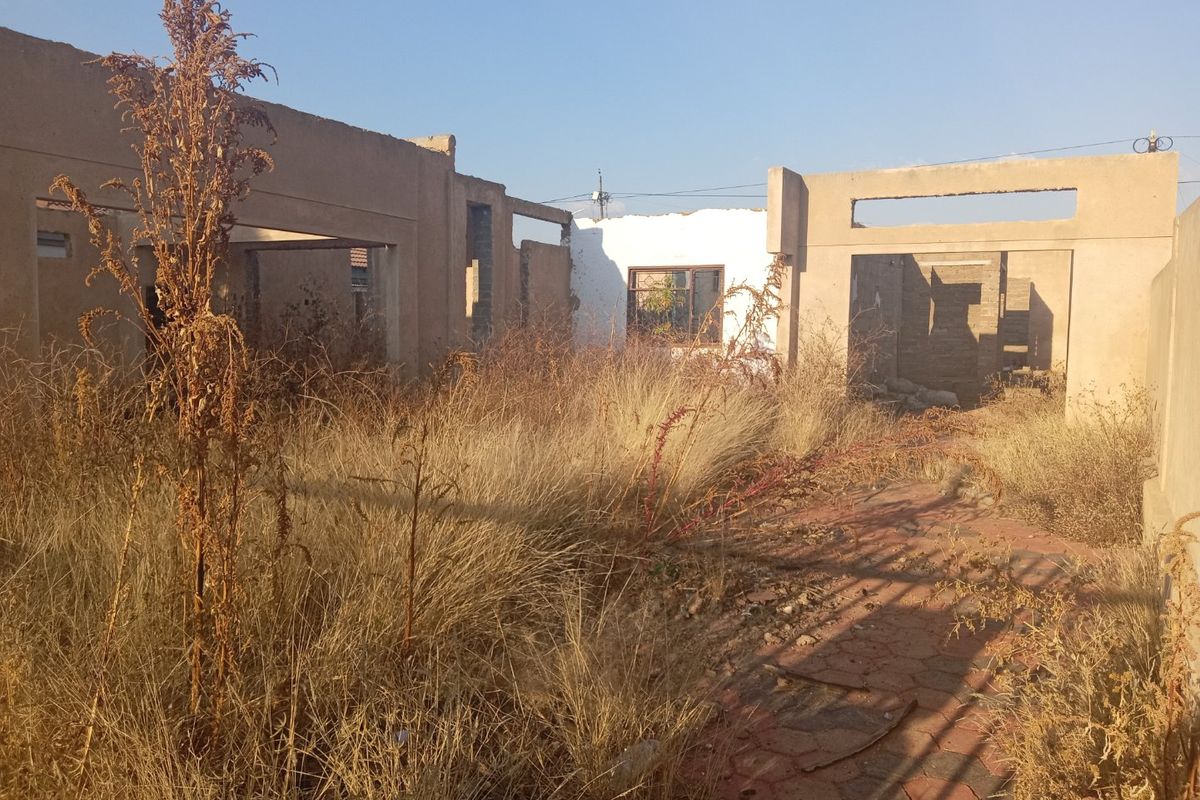 A view of a residential property under construction or in a state of disrepair, featuring exposed concrete walls and overgrown grounds.