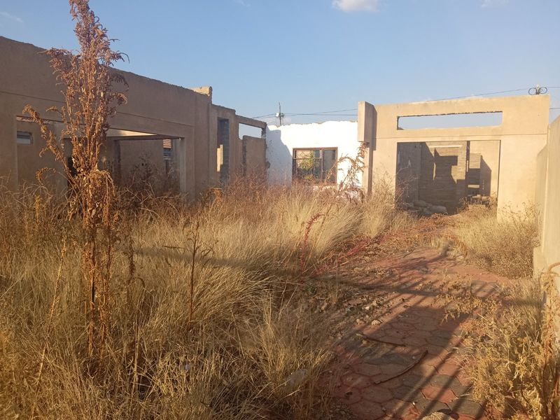 A view of a residential property under construction or in a state of disrepair, featuring exposed concrete walls and overgrown grounds.