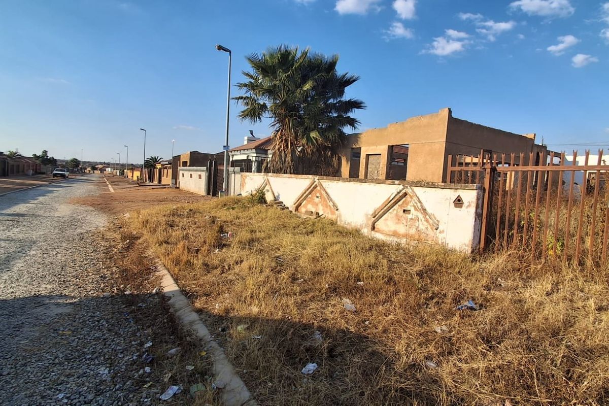 Vacant residential land featuring an unfinished building structure and a weathered boundary wall, situated along a gravel street.