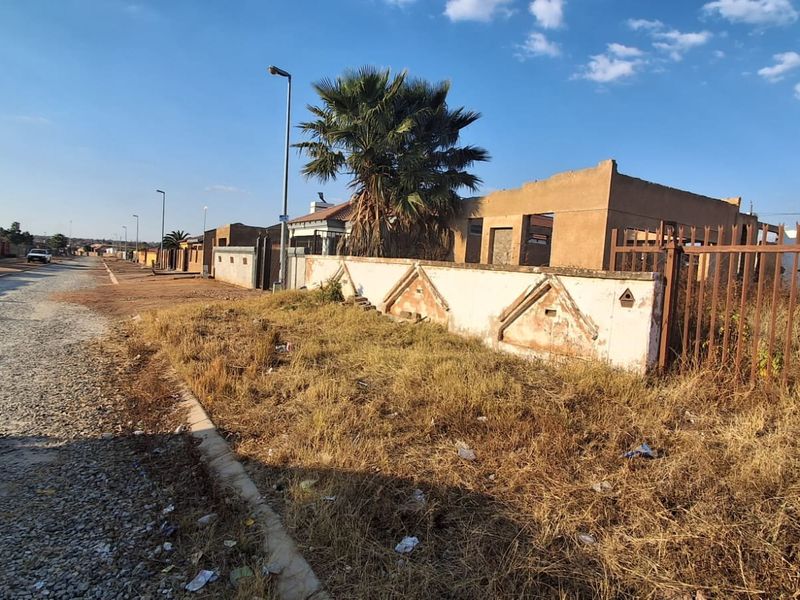 Vacant residential land featuring an unfinished building structure and a weathered boundary wall, situated along a gravel street.