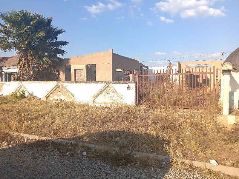 Street view of a vacant residential plot featuring an unfinished building structure, a weathered boundary wall, and a metal gate.