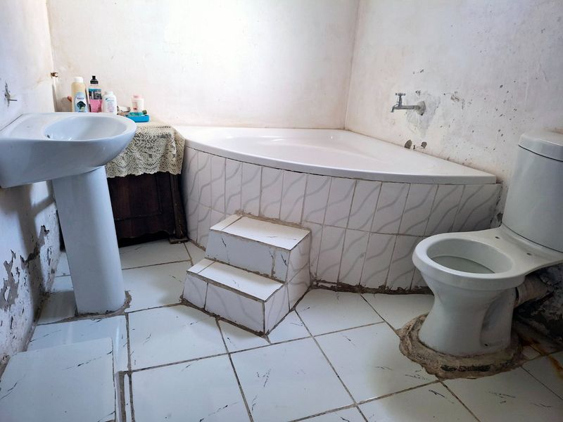 A dated bathroom featuring a corner bathtub with tiled steps, a pedestal basin, and a toilet, requiring significant cosmetic renovation and wall repairs.