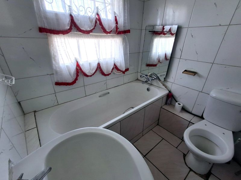 A dated bathroom featuring a built-in bathtub, toilet, and vanity basin with white ceramic wall and floor tiling.