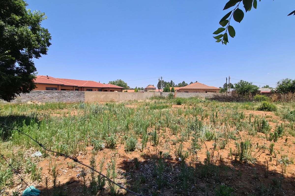 A vacant residential plot featuring natural ground cover, enclosed by a perimeter wall and wire fencing in a suburban setting.