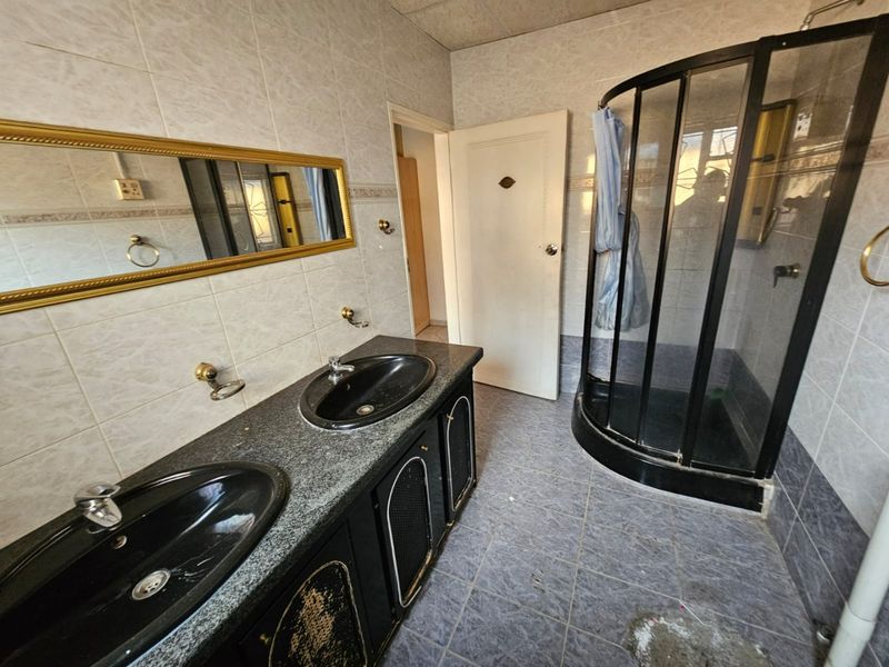 A dated bathroom featuring a double vanity with granite countertop, corner shower enclosure, and tiled finishes throughout, requiring cosmetic updates.