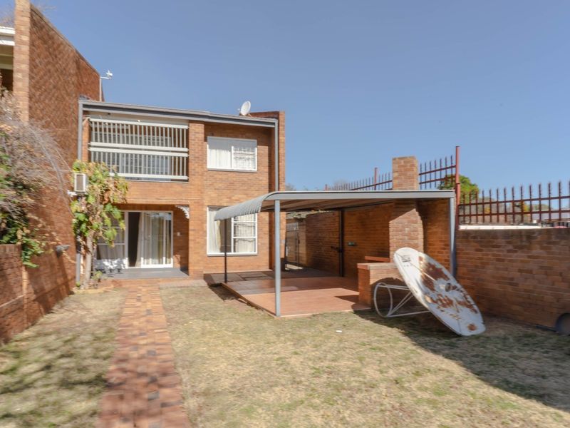 Rear view of a double-story brick townhouse featuring a covered patio area with a built-in braai, paved walkway, and enclosed garden space.