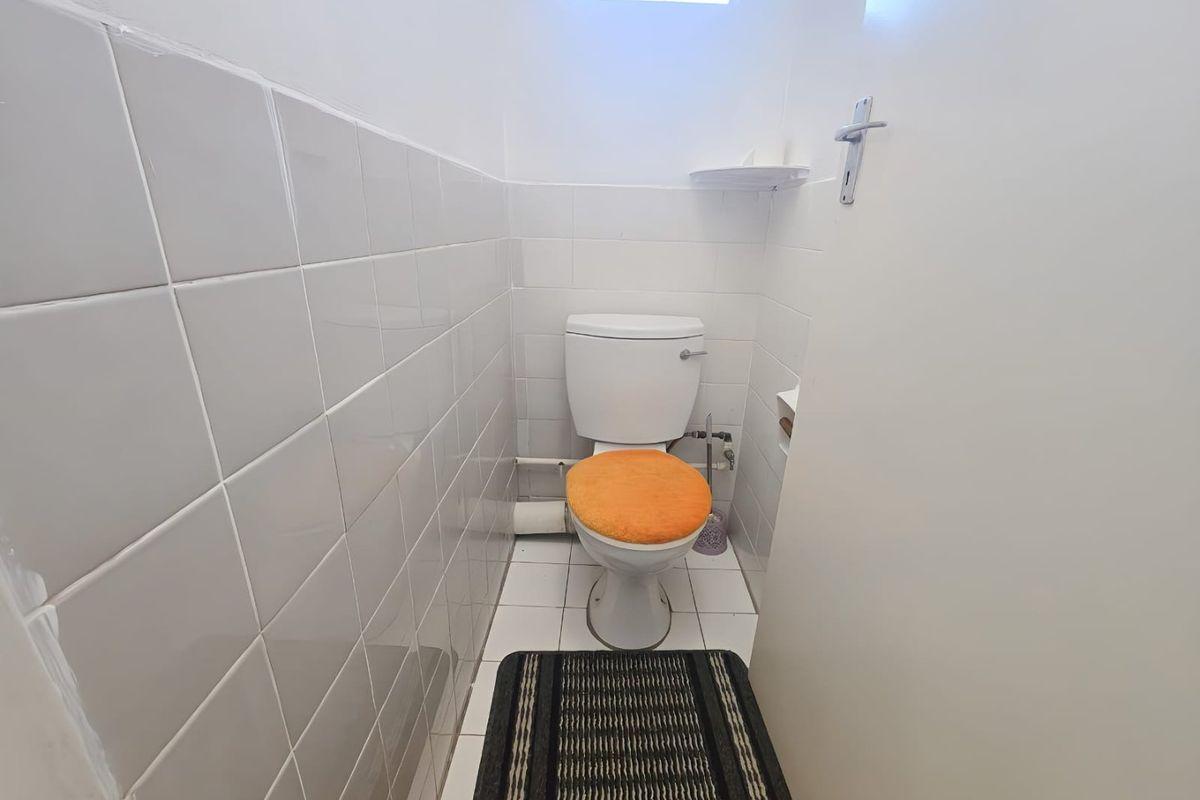A compact, functional bathroom featuring white ceramic wall and floor tiles, a standard toilet, and a small high-level window for ventilation.