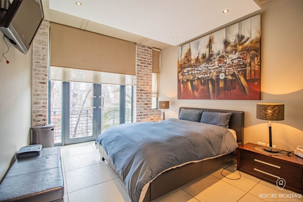 Modern bedroom featuring exposed brick accents, tiled flooring, recessed ceiling lights, and direct access to a private balcony through glass doors.