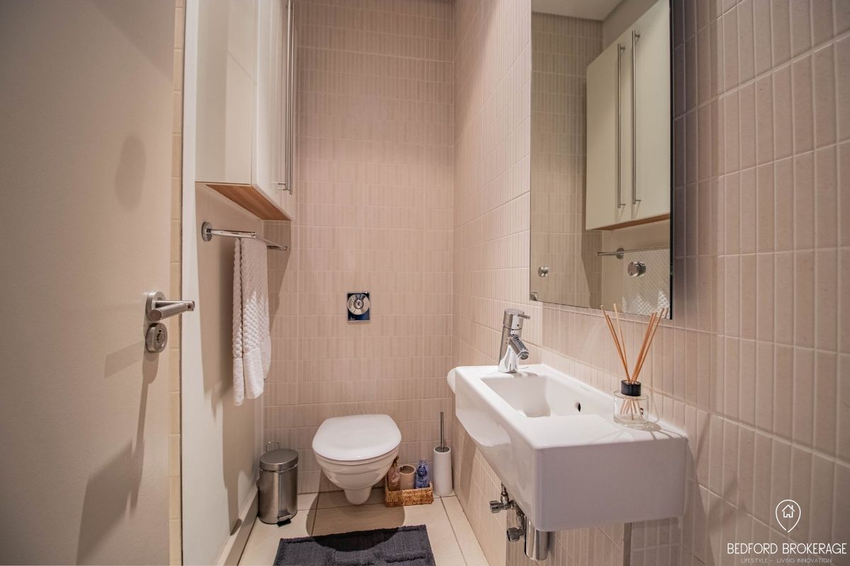 A modern, compact bathroom featuring a wall-mounted basin, wall-hung toilet, neutral tiled finishes, and integrated storage cabinetry.