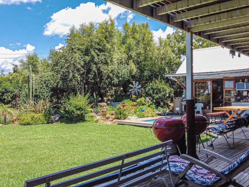 A covered wooden deck patio overlooking a lush garden and swimming pool area, providing a pleasant outdoor entertainment space.