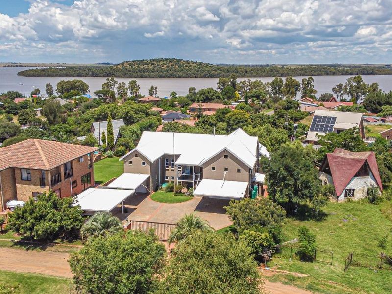 Aerial view of a residential property featuring a large house with a metal roof, paved driveway, and carports, situated near a lake.