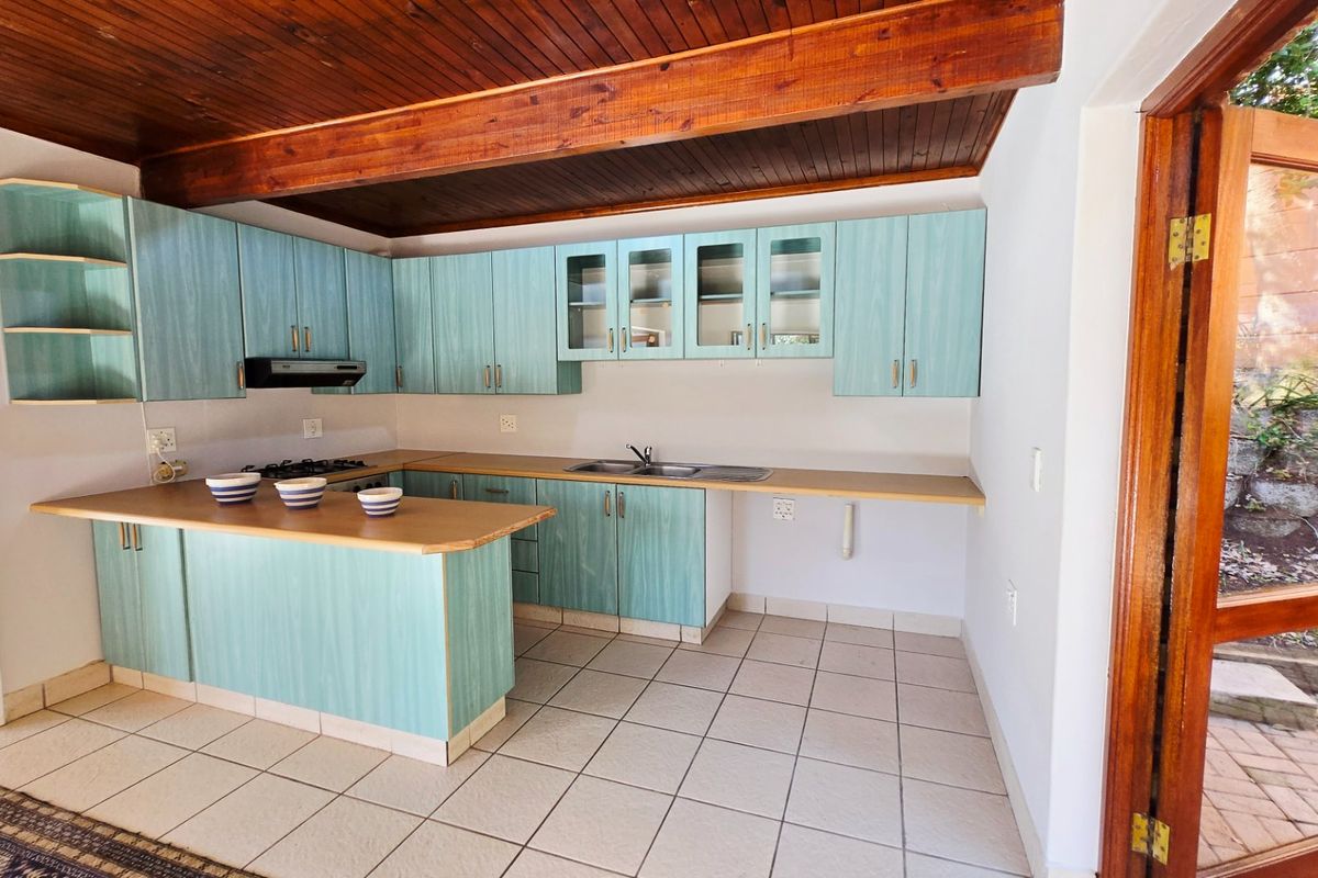 A functional kitchen featuring light blue laminate cabinetry, timber ceiling beams, and tiled flooring, with direct access to an outdoor area.