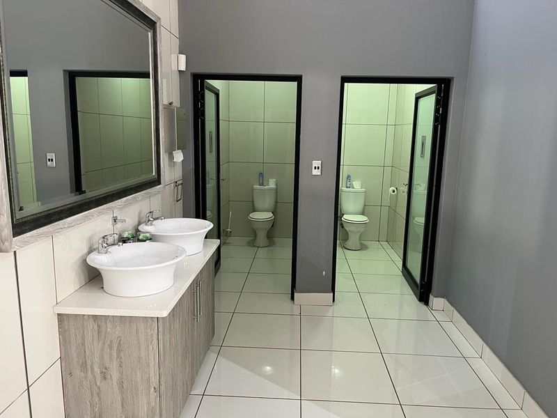 A clean, modern commercial bathroom featuring a dual vanity with vessel sinks, large wall mirror, and two separate toilet cubicles with tiled finishes.