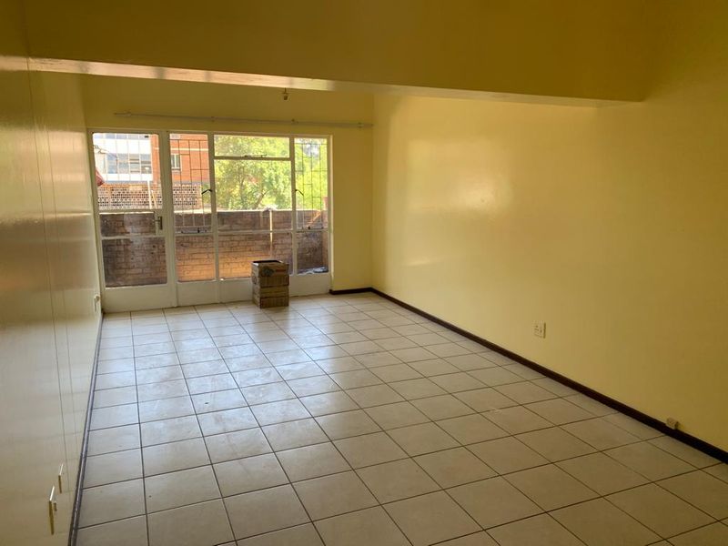 An empty, sunlit living room featuring neutral tiled flooring, built-in storage, and direct access to an outdoor balcony area through a sliding glass door.