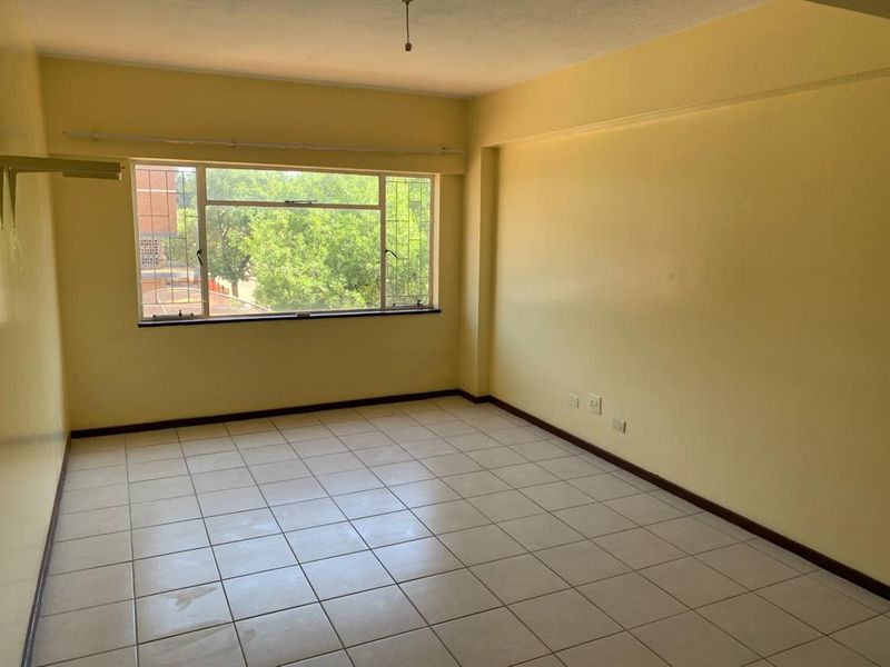 An empty, sunlit living room featuring neutral tiled flooring and a large window with security bars, offering a blank canvas for interior design.