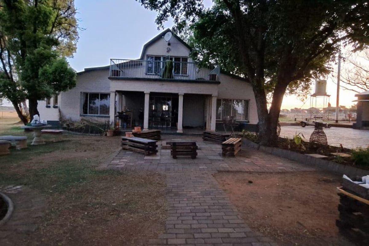 Rear view of the farmhouse featuring a covered veranda, upper-level balcony with metal railings, and a brick-paved patio area set within a garden.
