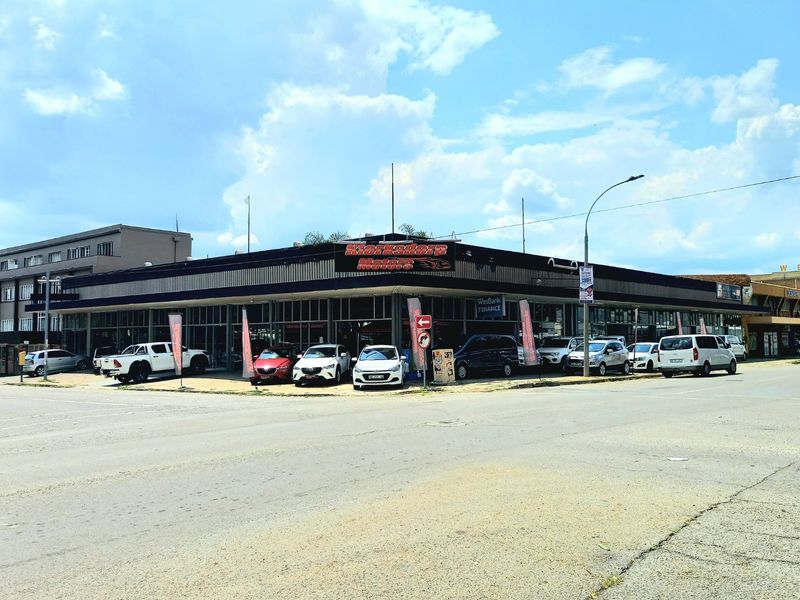 Commercial corner property featuring a large glass storefront, flat roof design, and paved forecourt area suitable for vehicle display or customer parking.