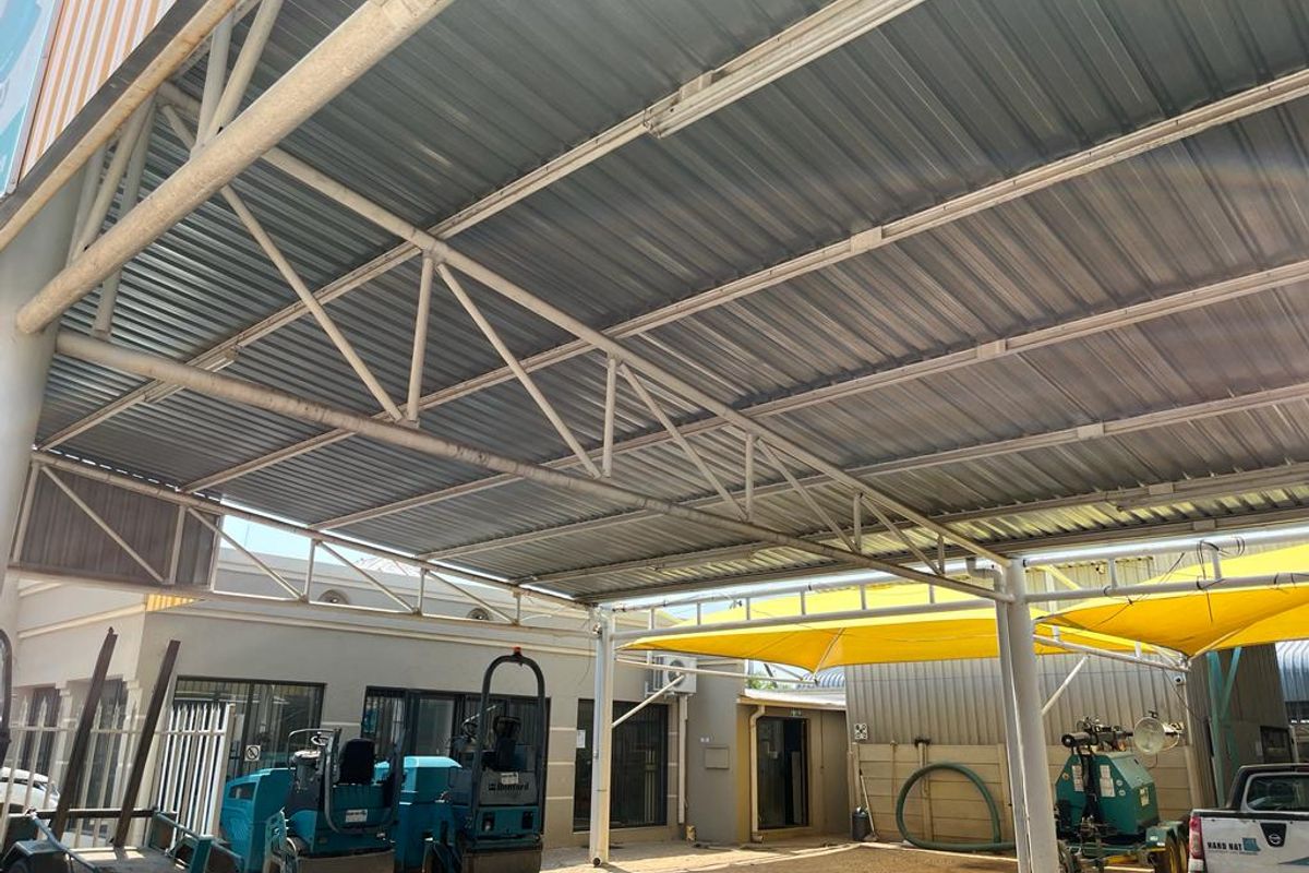 A commercial outdoor area featuring a large metal roof structure with steel trusses and shade sails, providing covered space for equipment or parking.