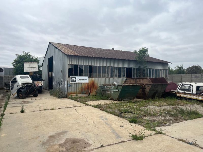 Industrial warehouse building featuring corrugated metal cladding, pitched roof, and extensive glazing, situated on a concrete hardstand area requiring maintenance.