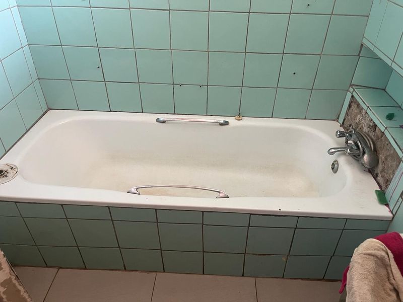 A dated bathroom featuring a built-in bathtub with chrome fixtures and light green wall tiling, requiring renovation to update finishes and address tile damage.