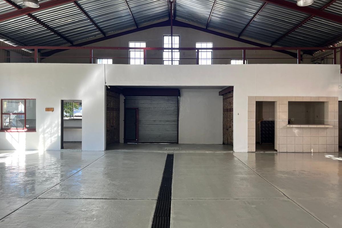 Spacious commercial utility or warehouse space featuring high ceilings with exposed steel trusses, concrete flooring, a roller shutter door, and a tiled service counter.