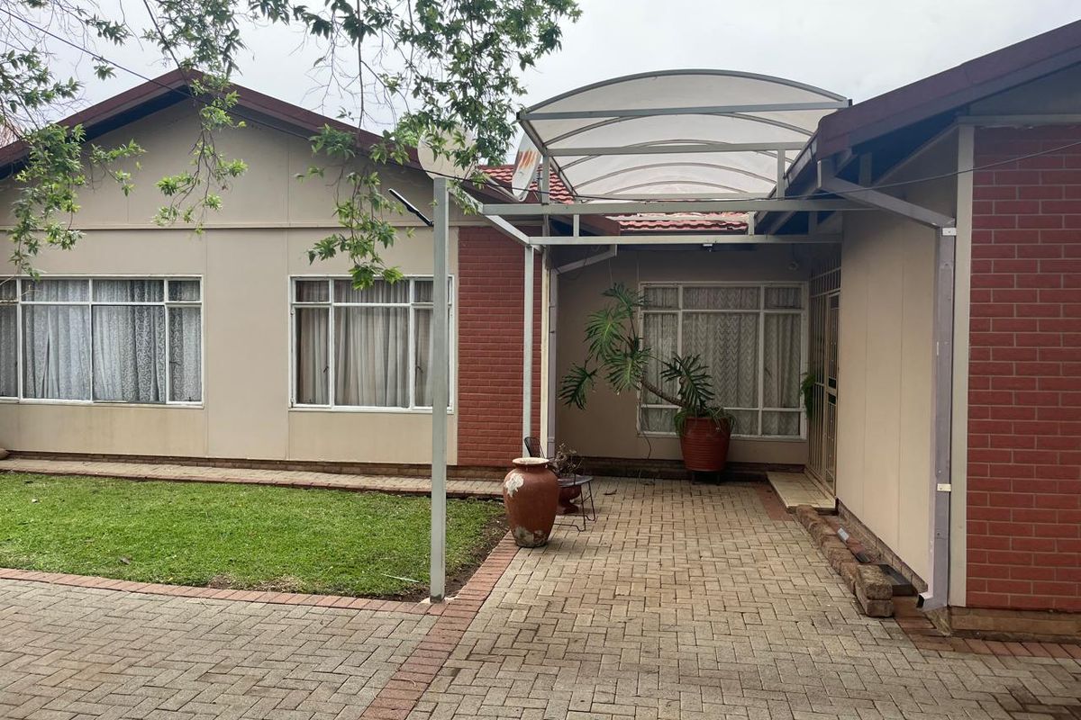 Rear exterior view featuring a paved patio area with a covered walkway, brick and plaster facade, and established garden space.