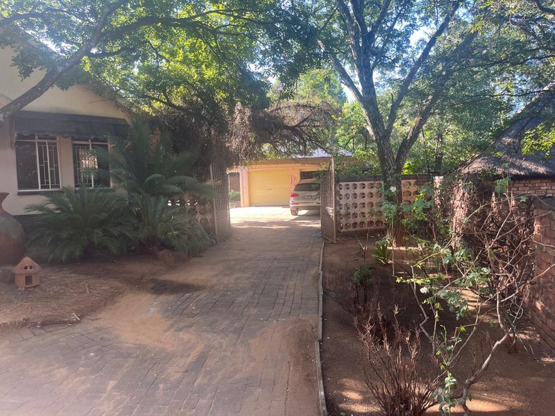 Front entrance view featuring a paved driveway, brick boundary wall, and access to a garage area surrounded by mature trees.