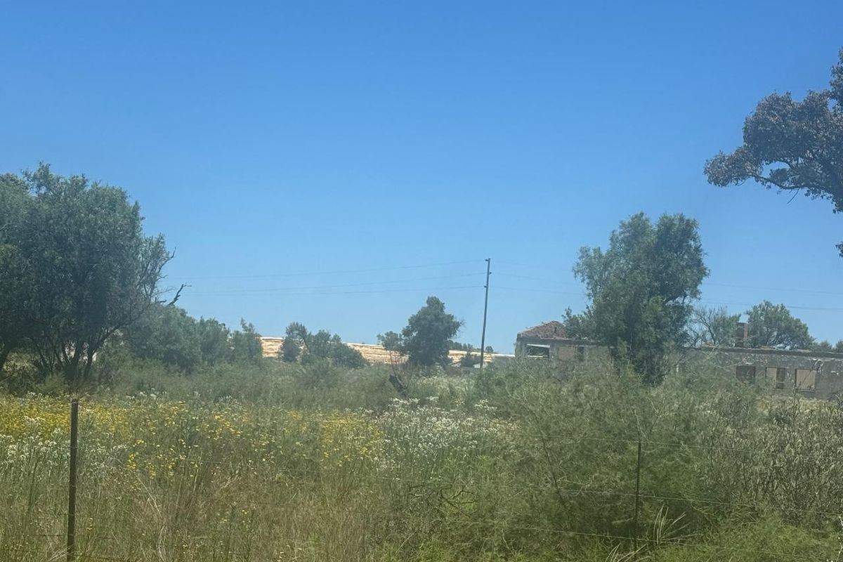 A view of a vacant residential plot featuring overgrown vegetation, wire fencing, and a dilapidated structure in the background.