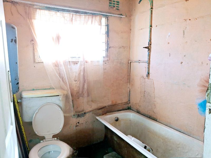 A dated bathroom requiring significant renovation, featuring a standard bathtub, toilet, and exposed plumbing with visible wall surface damage.