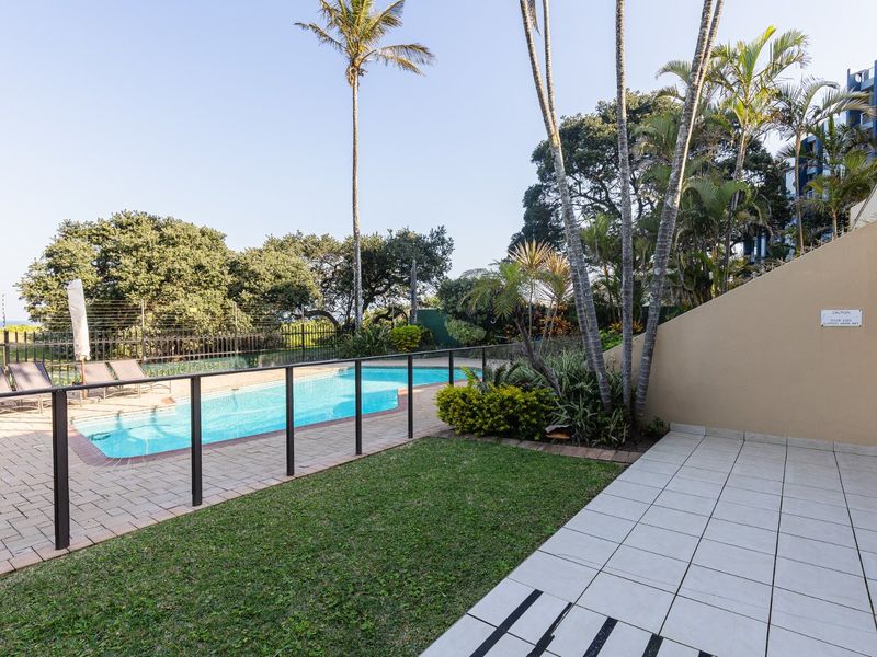 A well-maintained outdoor pool area featuring a paved deck, glass safety fencing, and a landscaped garden setting.